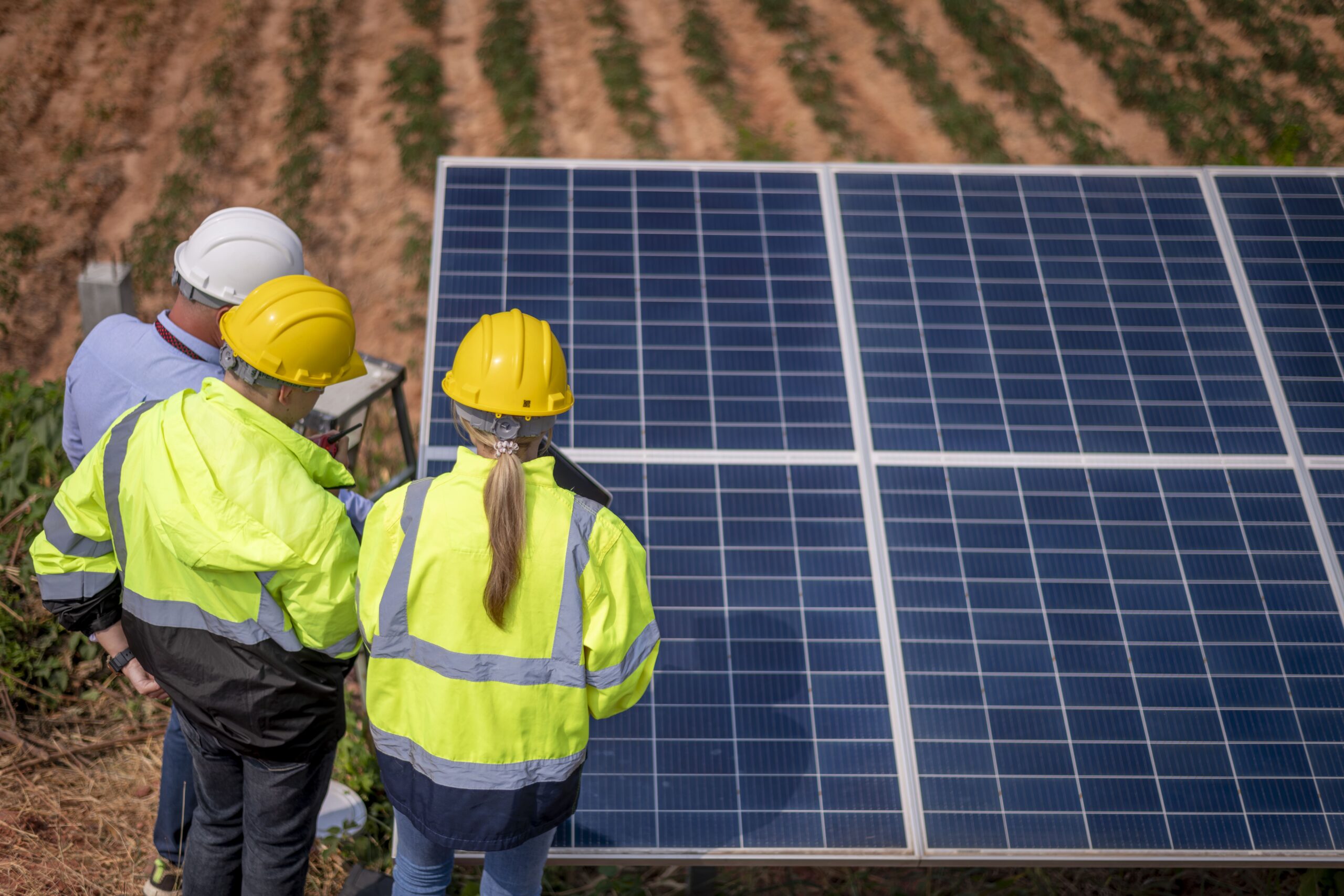 Dois engenheiros, um homem e uma mulher, com uniformes e capacetes amarelos, inspecionando um painel solar